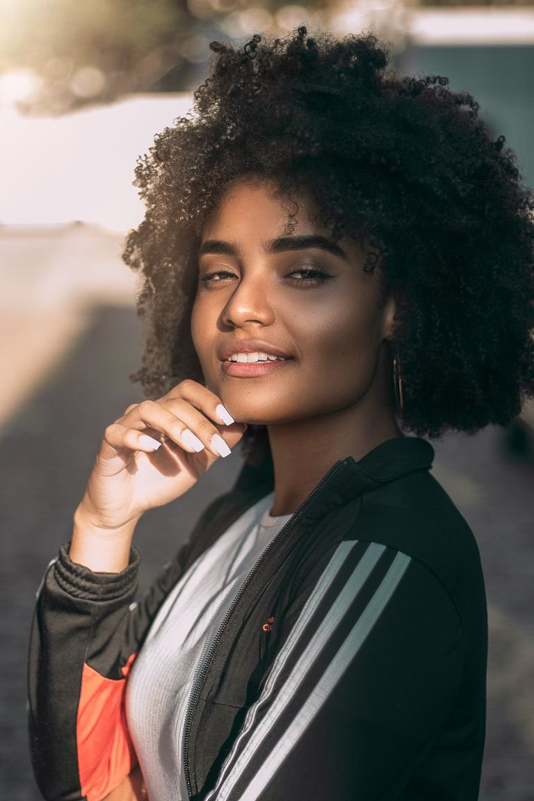 A beautiful woman with curly hair smiles warmly in a sunlit outdoor portrait.