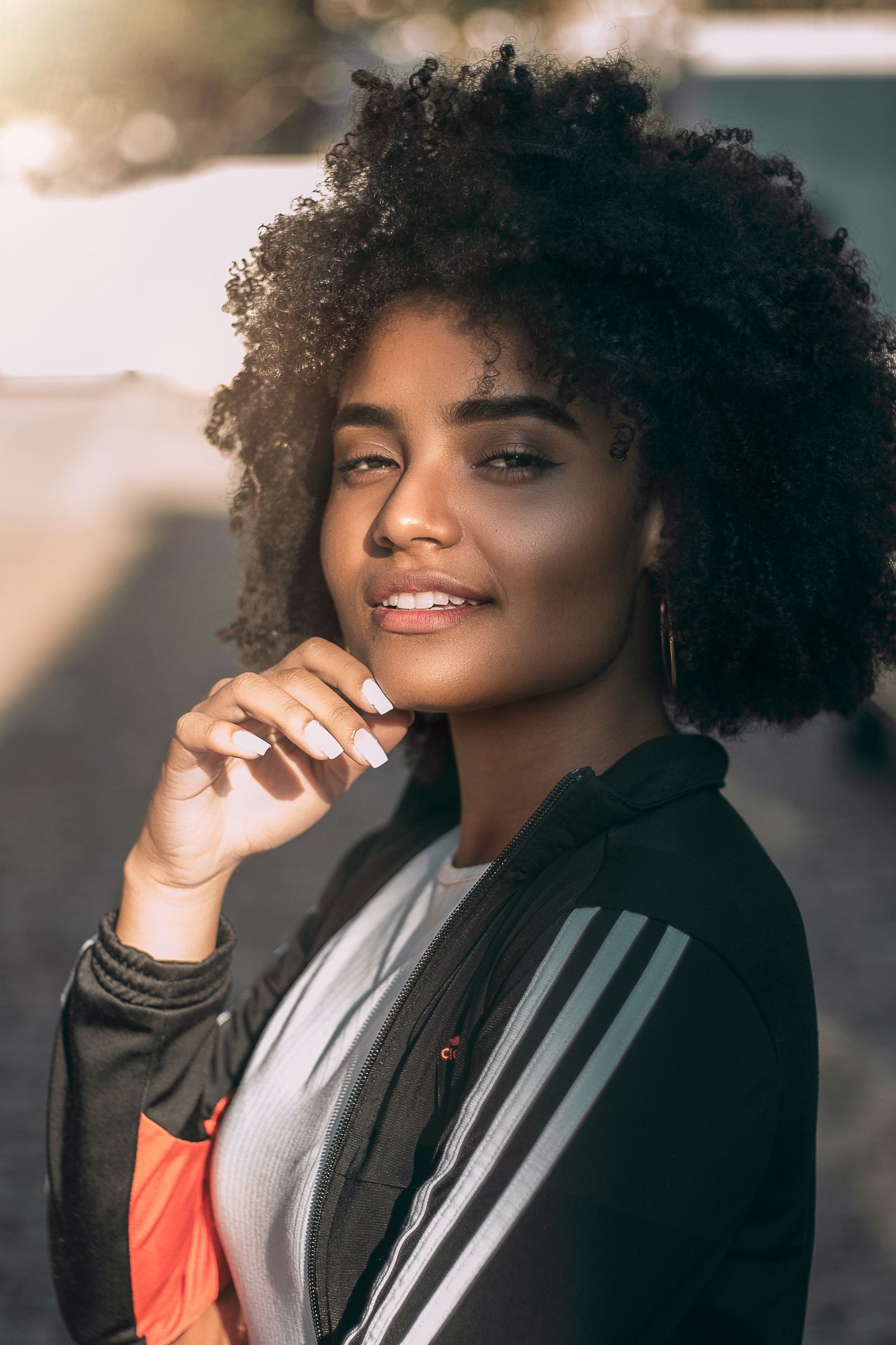 A beautiful woman with curly hair smiles warmly in a sunlit outdoor portrait.