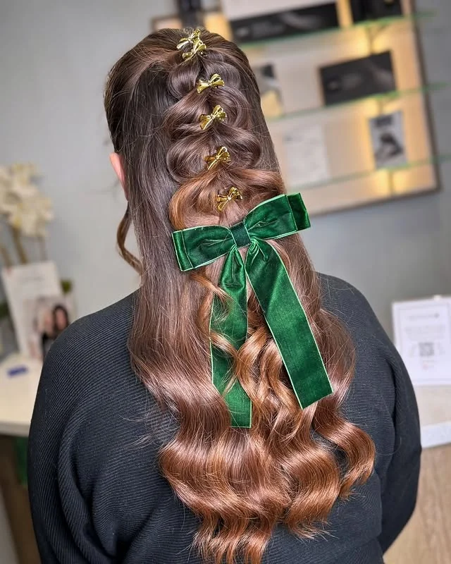 Back view of long wavy brown hair in a half-up style decorated with gold bow clips and a large green velvet bow.