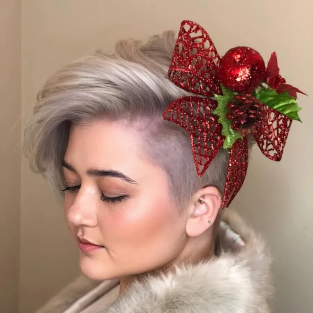 A woman with a platinum blonde pixie haircut wearing a glittery red Christmas bow clipped into the side.