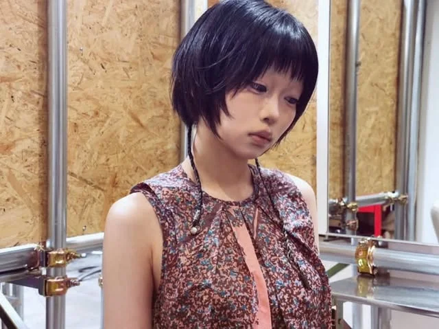 Front view of a woman with a short dark bob and blunt bangs, wearing two thin braided strands at the front.