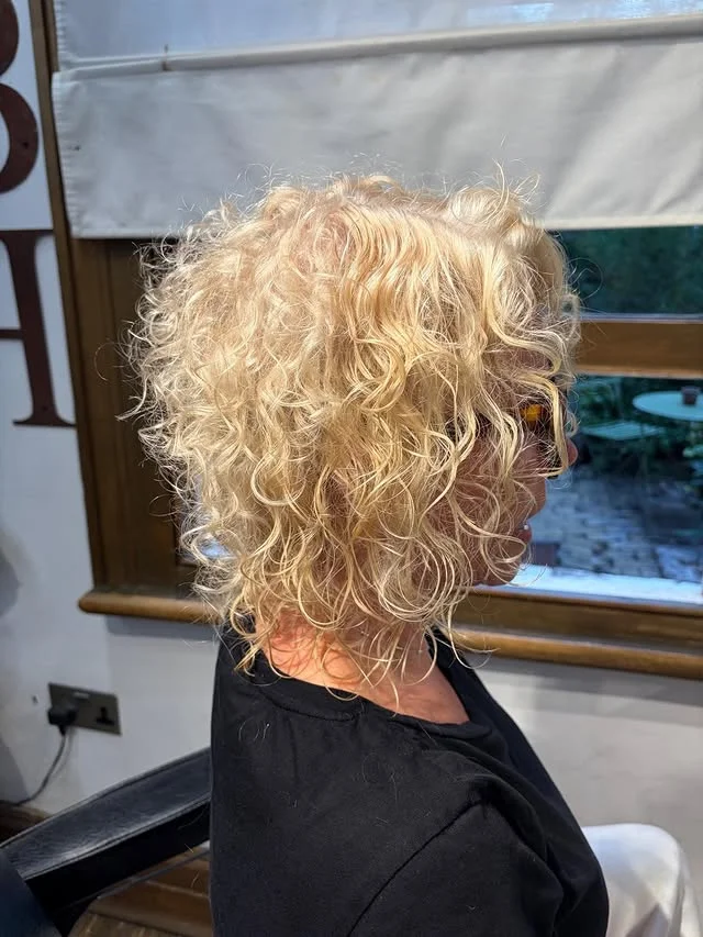 Rear view of a woman with a voluminous golden blonde curly bob in a salon.