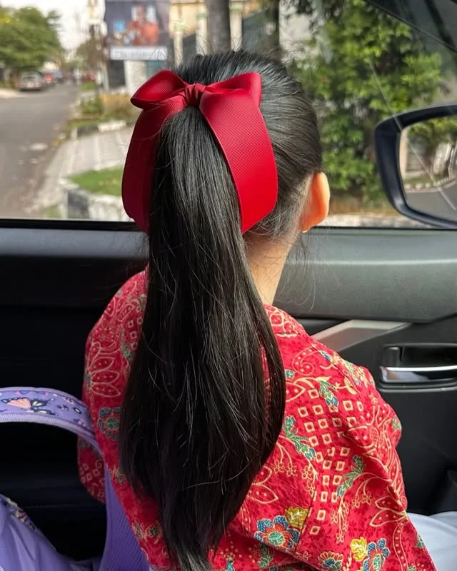 A young girl with long straight black hair in a high ponytail tied with a large red bow, seated in a car.
