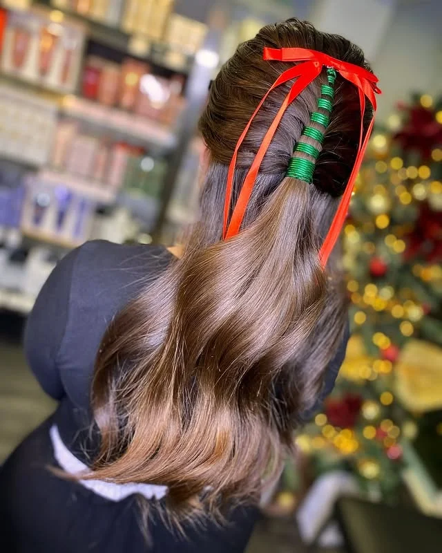 A brunette with a wavy low ponytail decorated with green braided bands and a large red satin bow, viewed from behind in a salon.