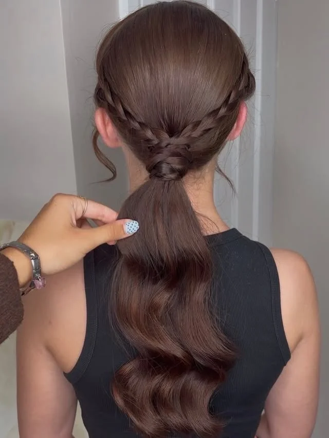 A woman with long dark wavy hair in a low ponytail with a braided section along the crown, photographed from behind in a salon.