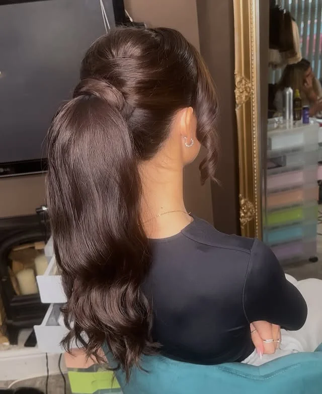 A woman with long dark brown wavy hair pulled into a voluminous high ponytail, seen from the side.