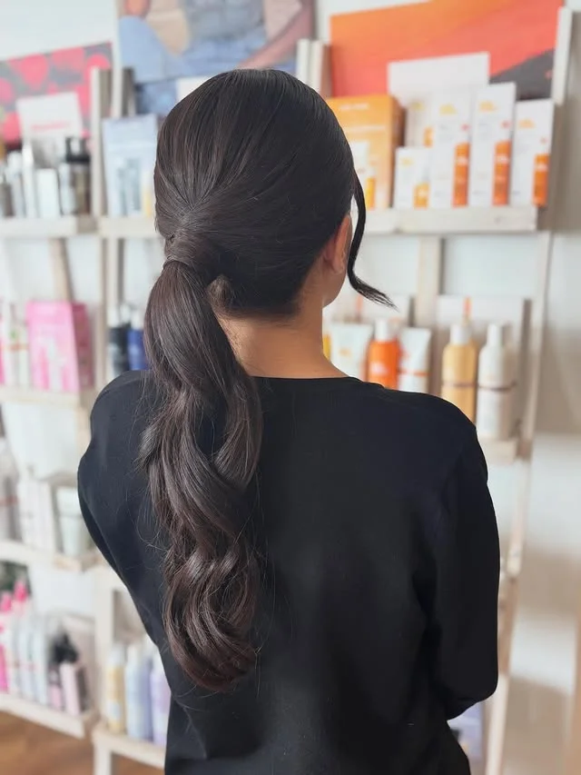 A woman with long dark wavy hair in a simple low ponytail, photographed from behind in a salon.