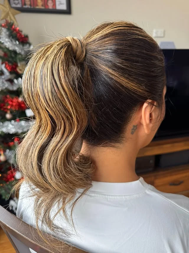 A woman with a high ponytail of soft blonde waves standing in front of a decorated Christmas tree.