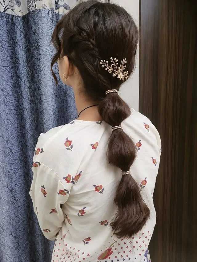 A woman with dark brown hair in a segmented bubble ponytail secured with pearl ties and a gold floral pin.