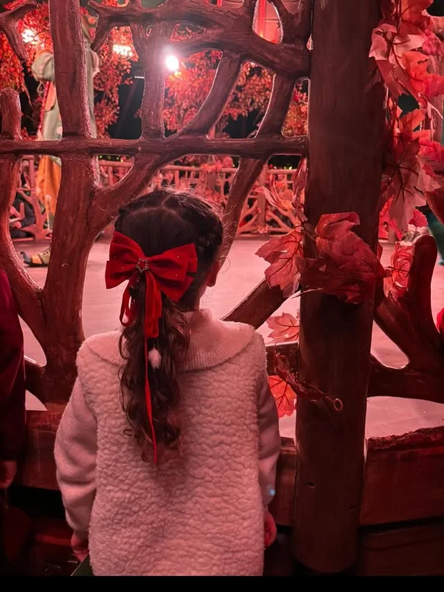 A young girl with dark curly hair pulled into a ponytail secured with a large red velvet bow with a jeweled center.