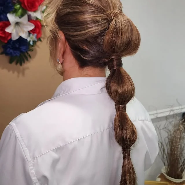 A woman with brown highlighted hair in a high bubble ponytail with hair wrapped sections, seen from behind.