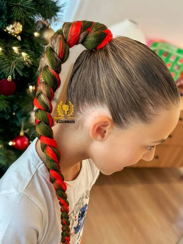A young girl with a high ponytail of red and green wrapped braids in front of a decorated Christmas tree.