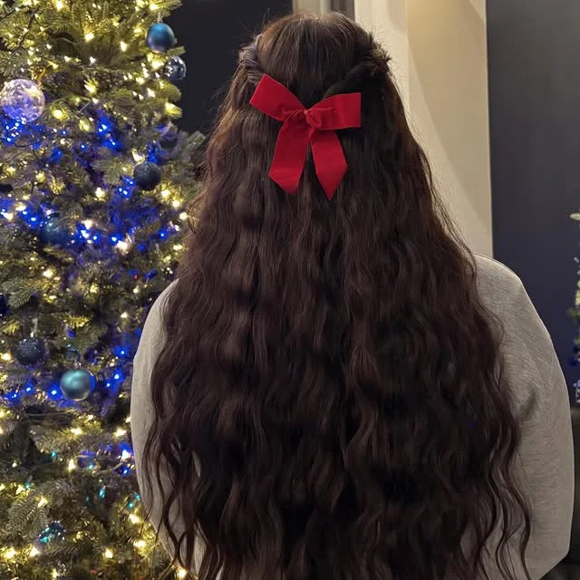 Rear view of long dark wavy hair with a red velvet bow clipped at the crown, beside a lit Christmas tree.
