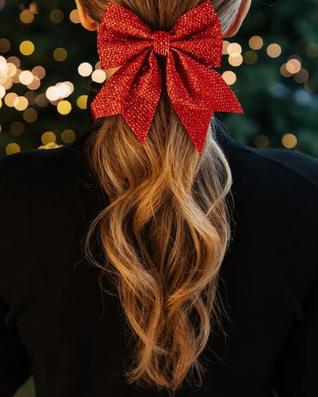 Sleek wavy low ponytail secured with an oversized glittery red bow, against a blurred Christmas tree.