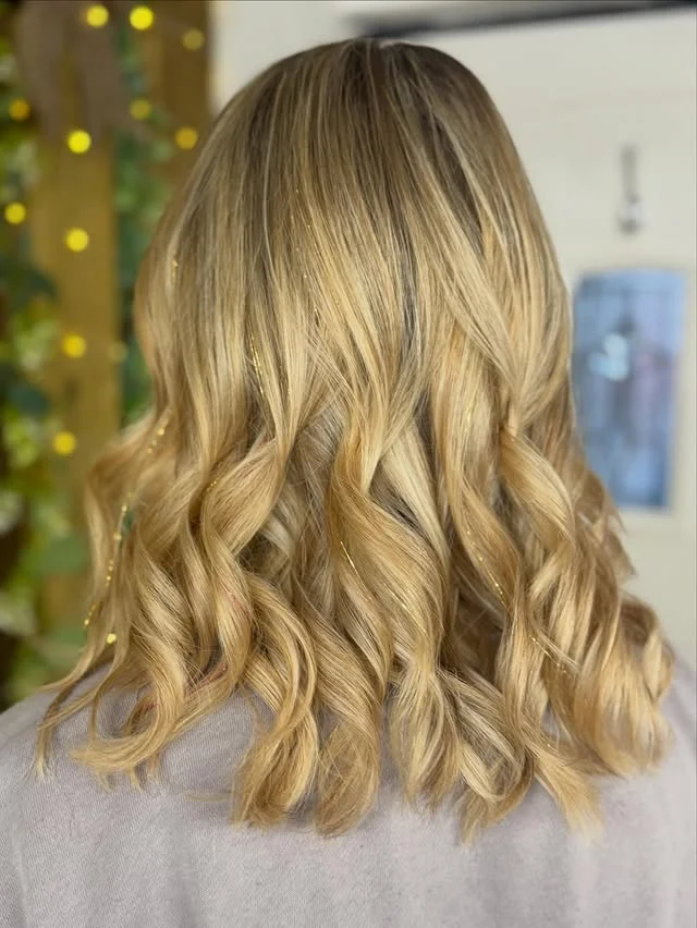 Back view of blonde wavy hair with thin gold tinsel strands in front of warm fairy lights.